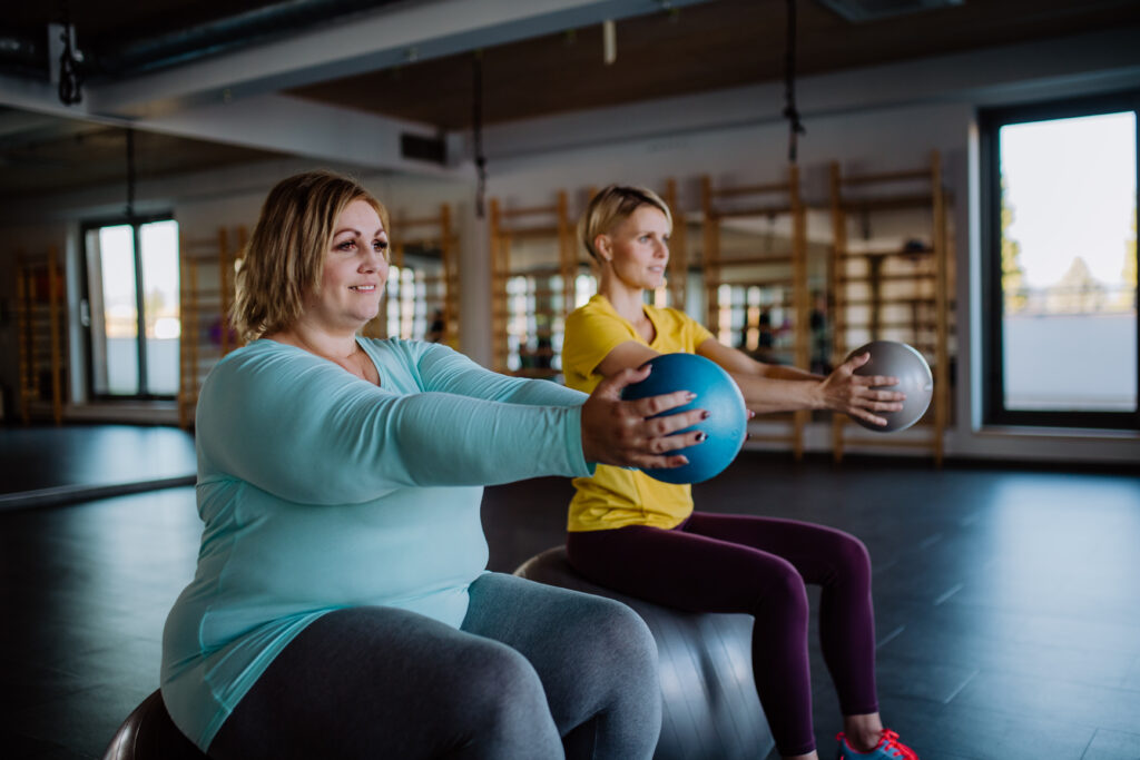 Happy overweight woman sitting on fintess ball and exercising with personal trainer in gym